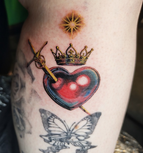 Tattoo eines Fantasy Motivs in Form eines Herzens, durchstochen mit einem Schwert, einer Krone, und eines Sterns auf dem Schienbein